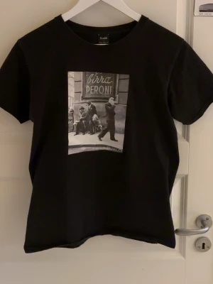 Svart t-shirt med print från Bvaldi - Svart t-shirt från Bvaldi med ett stort svartvitt print framtill som föreställer en stadsscen och texten 'Birra Peroni'. Klassisk rund halsringning och korta ärmar. Pris går att diskutera vid snabb affär😊