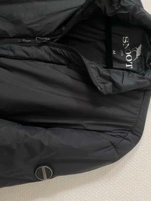 Svart pufferjacka från Snoot - Nypris 2000kr. Säljer en svart pufferjacka från Snoot med hög krage och dragkedja framtill. Bra skick förutom fickan på bröstet där dragkedjan är trasig (sista bilden) Jackan har två sidofickor med dragkedja och en diskret logotyp på ärmen. Tillverkad i ett glansigt, vindtätt material som håller dig varm.