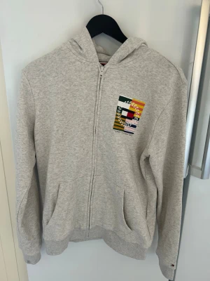 Grå hoodie från Tommy Hilfiger - Snygg ljusgrå hoodie med dragkedja från Tommy Hilfiger. Framtill finns ett färgglatt tryck med logga och texten 'Tommy Hilfiger 1985'. Hoodien har huva, två fickor och ribbade muddar. Perfekt för en avslappnad och trendig stil.