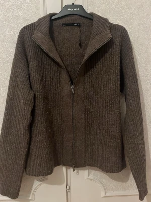 Brun stickad ziptröja från lager 157 - Mysig brun stickad tröja från lager 157 med dragkedja framtill och hög krage. Tröjan har ribbstickat mönster och långa ärmar, perfekt för kyliga dagar. Enkel och stilren design som passar till många outfits.