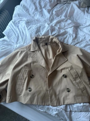 Kort beige trenchcoat med dubbelknäppning - Snygg kort trenchcoat i beige med klassisk dubbelknäppning och bred krage. Jackan har två framfickor 