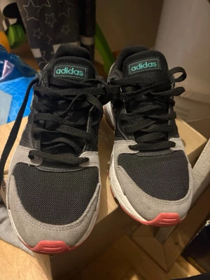 Adidas svarta och grå sneakers - Snygga Adidas sneakers i svart mesh och grå mocka med rosa detaljer på sulan. Skorna har klassisk låg profil, svart snörning och Adidas-logga på plösen i turkos. Perfekta för dig som gillar streetwear och sportig stil.
