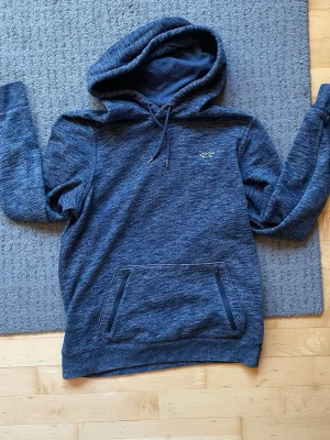 Mörkblå hoodie från Hollister - Snygg mörkblå hoodie från Hollister med broderad logga på bröstet och två dragkedjeförsedda fickor framtill. Tillverkad i mjukt, melerat material och har justerbar huva med dragsko. Perfekt för en avslappnad och cool stil.