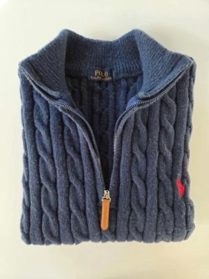 Mörkblå kabelstickad tröja Polo Ralph Lauren - Mörkblå kabelstickad tröja från Polo Ralph Lauren med quarter Zip och ribbad krage. Tröjan har det klassiska röda Polo-logot på bröstet och är tillverkad i mjukt, tjockt 100 % bomull. Perfekt för kalla dagar 
