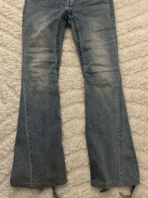 True Religion Joey Low Rise Flare Jeans - Säljer ett par blåa True Religion Joey jeans med låg midja och utsvängda ben. Klassisk tvätt med slitningar på låren och råa kanter nertill. Perfekta för dig som gillar vintagevibbar och en avslappnad look. Och det är uppsydda och om man spretar upp kommer det inte gå tillbaka till den ursprungliga längden