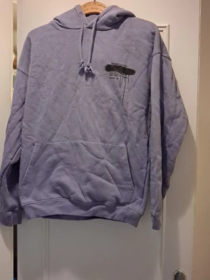 Ljuslila hoodie med tryck - Säljer en ljuslila hoodie med huva och känguruficka. På bröstet finns ett svart tryck med text och på ryggen står det 'RICH ONE DAY' i stor graffiti-stil. Hoodien har dragsko i halsen och är i mjukt material, perfekt för en avslappnad streetwear-look.