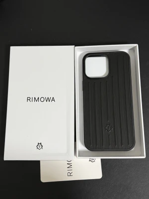 Svart iPhone 16 Pro Max mobilskal från Rimowa - Snyggt svart mobilskal från Rimowa med räfflad design och diskret logga på baksidan. Skalet är tillverkat i ett tåligt material och har en stor öppning för kameran. Perfekt för dig som vill ha ett stilrent och exklusivt skydd till din mobil. För 16 pro max