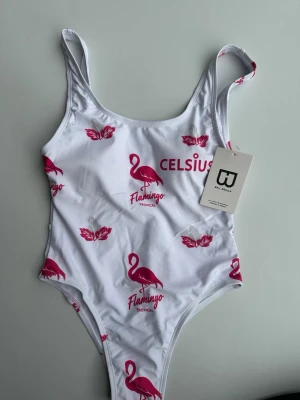 Vit baddräkt med flamingotryck - Snygg vit baddräkt från Ball Bralla med rosa flamingos, blad och texten 'CELSIUS Flamingo Tropical' tryckt framtill. Klassisk modell med smala axelband och hög skärning. Perfekt för strandhäng eller poolparty.