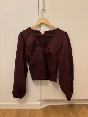 Vinröd satinblus med v-ringning - Supersnygg vinröd blus i satin med v-ringning och smockad midja. Blusen har långa puffärmar och en figurnära passform som framhäver midjan. Perfekt att styla med både jeans och kjol för en trendig look. Slutsåld på Gina, jag har använt den en gång. Den är i nyskick 