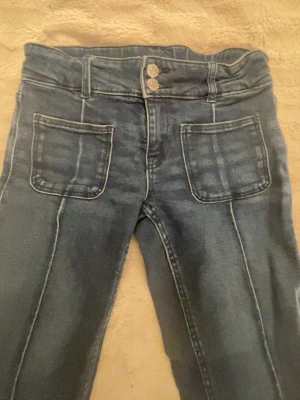 Blå jeans med dubbla knappar - Säljer ett par blå jeans med raka ben och dubbla knappar i midjan. Jeansen har stora framfickor och markerade sömmar längs benen. Klassisk tvättad denim-look som passar till det mesta. Materialet är troligtvis bomull med stretch.