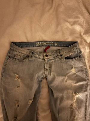 Ljusblå slitna jeans från Guess-Reserverad  - Säljer ett par ljusblå jeans från Guess med slitningar och distressed detaljer på både fram- och baksida. Klassisk femficksmodell med normal midja och knappgylf. Snygg broderad bakficka och diskret logga🩵