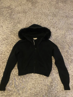 14+ Fur Hoodie - Svart - Säljer en svart stickad fur hoodie från Gina Tricot med dragkedja och huva som har en snygg kant av fuskpäls. Tröjan är alldrig använd, ligger kvar i förpackningen, helt nytt skick med prislapp kvar.  Säljer den för att den inte kommer till användning.Tröjan har ribbade muddar vid ärmslut, midja, och en croppad passform. Perfekt för dig som gillar en trendig och mysig look. 💕