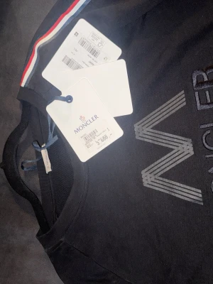 Svart Moncler sweatshirt med ränder - (KÖPT PÅ SICURO FASHION) Säljer en svart sweatshirt från Moncler med diskret logga på bröstet och vita/röda ränder längs ärmarna. Tröjan har rund halsringning och långa ärmar. Perfekt för dig som gillar stilrena och sportiga plagg.