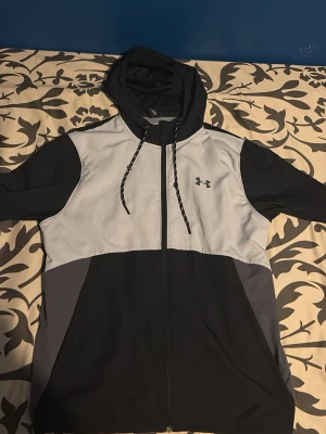 Svart och grå vindjacka Under Armour - Snygg vindjacka från Under Armour i svart, grått och ljusgrått. Jackan har huva med dragsko, hel dragkedja framtill och två sidofickor. Tillverkad i lätt och vindtätt material med meshfoder för extra komfort. Perfekt för blåsiga dagar.