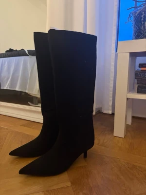 NYA Svarta stövlar med klack storlek 38 - Säljer ett par helt oanvända svarta knähöga stövlar med smal spetsig tå och låg klack. Klackar är köpta från Nelly, är helt oanvända och ligger i kartong. Stövlarna har en elegant siluett och är tillverkade i ett mjukt material som ser ut som mocka. Perfekta för att lyfta vilken outfit som helst. Nypris 800kr jag säljer för 600