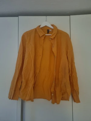 Gul långärmad linneskjorta - Snygg orange skjorta i linne med klassisk krage och knappar framtill. Skjortan har lång ärm och en avslappnad passform, perfekt för dig som gillar en enkel men färgstark stil soppar uppmärksamhet. Passar lika bra till jeans som till shorts.