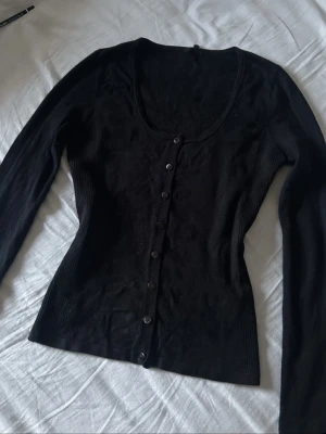 Svart ribbad kofta med knappar - Snygg svart ribbad kofta med långa ärmar och rundad urringning. Koftan har en figurnära passform och knäpps med svarta knappar framtill. Perfekt att styla över ett linne eller t-shirt för en enkel men trendig look.