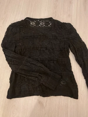 Svart spetsblus med volangdetaljer - Snygg svart blus i spets med blommönster och volangdetaljer över hela plagget. Transparent design som ger en trendig look. Rund halsringning och långa ärmar. Perfekt att styla sopistikerat eller edgy beroende på humör.