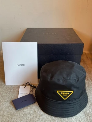 Svart bucket hat från Prada - Snygg svart bucket hat från Prada med det klassiska triangulära logomärket i gult och svart framtill. Använd ett fåtal gånger!