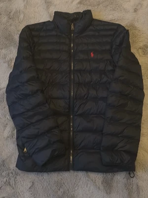 Mörkblå pufferjacka från Ralph Lauren - Snygg Mörkblå pufferjacka från Ralph Lauren med diskret röd logga på bröstet. Jackan har hög krage, dragkedja framtill och quiltad design. Perfekt för kalla dagar och har en klassisk, stilren look som funkar till det mesta.