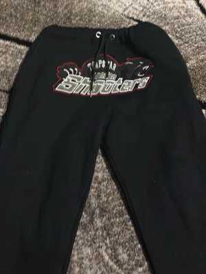 Svart hoodie Trapstar Shooters - Svart hoodie från Trapstar med stort Shooters-tryck på bröstet och coola detaljer i vitt och rött. Klassisk huva med snören och mjukt material som känns skönt mot huden. Perfekt för dig som gillar streetwear och vill sticka ut.