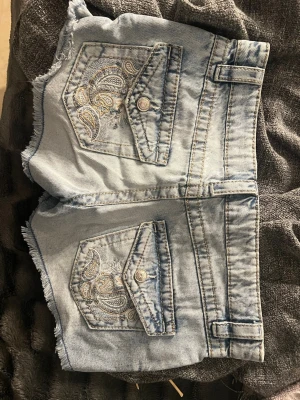 Ljusblå denimshorts med broderi - Säljer ett par ljusblå denimshorts med råa kanter och snygga broderade detaljer på bakfickorna. Shortsen har fickor både fram och bak, bälteshällor och knappgylf. Perfekta för sommaren och ger en avslappnad vibe.