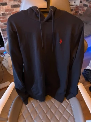 Svart hoodie från US polo assn - Svart hoodie från US polo assn med klassisk huva och dragsnören. På bröstet finns den ikoniska röda Polo-loggan broderad. Hoodien har känguruficka framtill och är tillverkad i mjukt bomullsmaterial. Perfekt för en chill och stilren look.