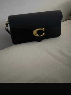 Svart kuvertväska från Coach - Snygg svart kuvertväska från Coach i läder med guldfärgade metalldetaljer och ett singular C-spänne framtill. Väskan har en avtagbar axelrem och en klassisk Coach-tag i läder. Perfekt för dig som gillar stilrena accessoarer.