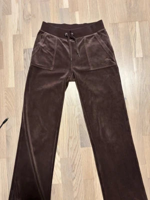 Bruna mjukisbyxor från Juicy Couture - Mörkbruna mjukisbyxor  från Juicy Couture med bred resårmidja och dragsko. Byxorna har två stora framfickor och två bakfickor med knappdetaljer. Perfekta för en chill och avslappnad stil.