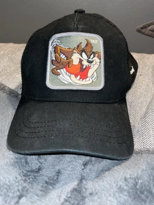 Svart Capslab keps med Taz-motiv - Snygg svart keps från Capslab med mesh på sidorna och ett stort broderat Taz-motiv från Looney Tunes framtill. Justerbar baktill för perfekt passform. Perfekt för dig som-vill sticka ut med en cool och lekfull accessoar.