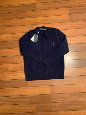 Mörkblå kabelstickad tröja Polo Ralph Lauren - Mörkblå kabelstickad tröja från Polo Ralph Lauren med rund halsringning och röd broderad logga på bröstet. Tröjan har ribbade muddar vid ärmslut och nederkant. Klassisk och tidlös design i mjukt material.