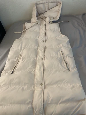 Ljusbeige lång dunväst med huva - Snygg ljusbeige lång dunväst med huva och dragsko. Västen har två fickor med dragkedja och stängs med både dragkedja och tryckknappar. Perfekt för lager på lager och ger en clean look till din outfit.