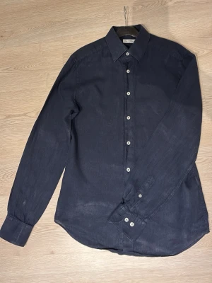 Mörkblå linneskjorta från these glory days - Stilren mörkblå skjorta i linne med klassisk krage och vita knappar framtill. Skjortan har lång ärm och en avslappnad passform, perfekt för en clean och enkel look.