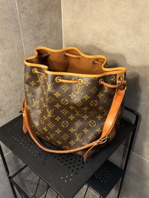 Louis Vuitton Noé - Louis Vuitton Noé med det klassiska monogramet. Använd men väl omhändertagen. Överlag väldigt fint skick, inga defekter som märks av vid använding. Bandet har några små slitningar och därför förstärkts med extra läder på insidan (se bild 6 och 7), syns ej vid använding. Självklart äkta. För fler bilder eller funderingar, skriv till mig privat!