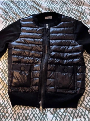 Svart pufferjacka från Moncler - Snygg svart pufferjacka från Moncler med quiltad front i glansigt material och stickade ärmar. Två stora fickor framtill med knappstängning och dragkedja hela vägen. Klassisk Moncler-logga på ärmen. Perfekt för kalla vinterdagar.