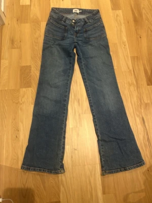 Blå bootcut jeans med låg midja - Ett par klassiska blå bootcut jeans med låg midja och utsvängda ben. Jeansen har framfickor med knappdetaljer och två bakfickor. Tillverkade i slitstarkt denimtyg med synliga sömmar i kontrastfärg. Perfekta för en avslappnad och trendig look.