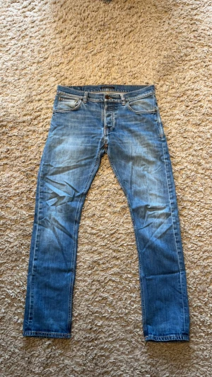 Blå jeans från Nudie Jeans - Säljer ett par klassiska blå jeans från Nudie Jeans med snygg slitning och raka ben. Jeansen har fem fickor, orange sömmar och Nudie-logga på bakfickan. Tillverkade i denim av bomull och har en tidlös look som passar till det mesta. Sjukt fräsch tvätt. Storlek 32/32