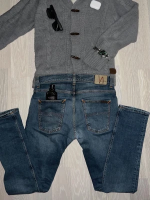 Blå jeans från Nudie Jeans - Säljer ett par klassiska blå jeans från Nudie Jeans med raka ben och femficksmodell. Jeansen har snygga kontrastsömmar och en patch med Nudie-loggan bak i midjan. Perfekta till vardagslooken och tillverkade i slitstark denim. Jenasen är lagade i skrevt snyggt och proffsigt av nudie så syns knappt när dom är på