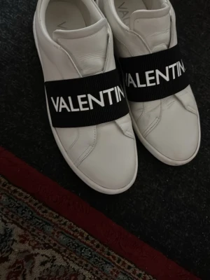 Vita Valentino sneakers med bred rem - Stilrena vita sneakers från Valentino med bred svart elastisk rem över foten där märkesnamnet står i vitt. Skorna är låga och har en minimalistisk design i slätt läder. Perfekta för dig som gillar exklusiva och trendiga detaljer.