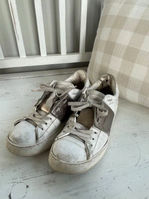 Vita sneakers med metallic-detaljer - Snygga vita Valentino sneakers med breda brons detaljer på sidorna och gråvita skosnören. Klassisk låg modell med rund tå och vit sula. 