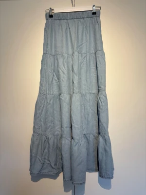 Ljusblå volangkjol med resårmidja - Säljer en ljusblå långkjol med volanger och resår i midjan. Kjolen har en avslappnad passform och är tillverkad i ett mjukt bomullsliknande material. Perfekt för dig som gillar en enkel men snygg stil.