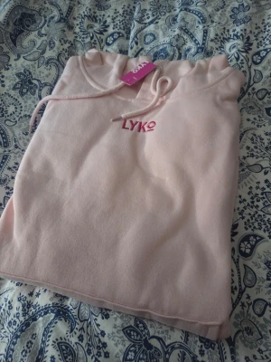 Ljusrosa hoodie från Lyko - Säljer en ljusrosa hoodie från Lyko med huva och dragsko. Framtill finns en liten broderad Lyko-logga och på ryggen en stor tryckt logga i rosa nyanser. Hoodien är i mjukt material, perfekt för en chill och avslappnad stil.