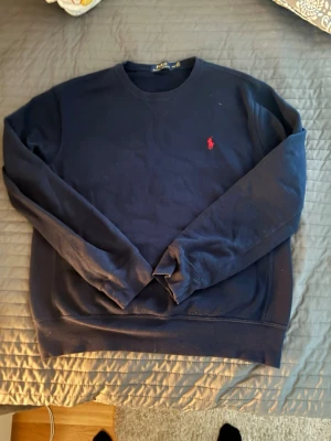 Mörkblå sweatshirt från Polo Ralph Lauren - Klassisk mörkblå sweatshirt från Polo Ralph Lauren med rund sopringning och den ikoniska röda logotypen broderad på bröstet. Tröjan har långa ärmar och ribbade muddar vid ärmslut och nederkant. Perfekt för en avslappnad exposure.