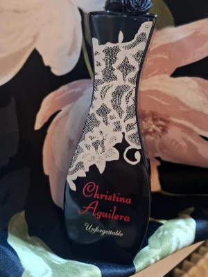 Christina Aguilera Unforgettable parfym - Elegant parfymflaska i svart glas med vit spetsliknande dekor och röd text. Flaskan har en kurvig form och ett svart lock med blomdetalj. Doften är från Christina Aguilera och heter Unforgettable, perfekt för dig som vill sticka ut.