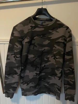 Camo-mönstrad sweatshirt från H&M - Cool sweatshirt med camo-mönster i svart, grått och mörkgrönt från H&M. Tröjan har rund halsringning och långa ärmar, perfekt för en avslappnad streetstyle-look. Priset kan diskuteras vid snabb affär.