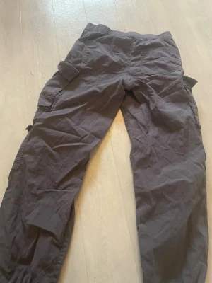 Mörkbruna cargobyxor med fickor - Säljer ett par mörkbruna cargobyxor med flera stora fickor på sidorna och dragsko vid bensluten. Byxorna har en avslappnad passform och är tillverkade i ett mjukt bomullstyg. Perfekta för en chill och street-inspirerad stil.
