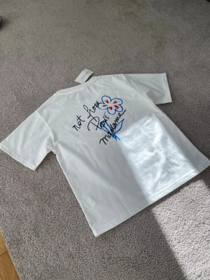 Drole De Monsieur Not From Paris Madame tryck - Vit t-shirt från Not From Paris Madame med färgglatt tryck av en blomma och text på bröstet och ryggen. Klassisk passform med rund hals och korta ärmar. Tillverkad i mjuk bomull, perfekt för en avslappnad och trendig look.