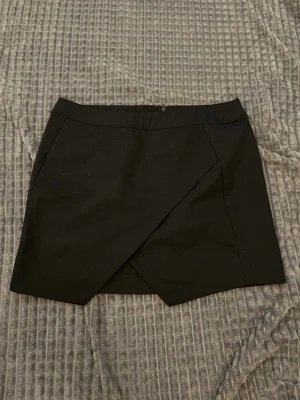 Svart omlottkjol i kort modell - Stilren svart kortkjol med omlott-design framtill. Kjolen har en rak passform och dragkedja bak. Perfekt att matcha med en snygg topp eller stickad tröja. Enkel och tidlös design som passar till många olika stilar.