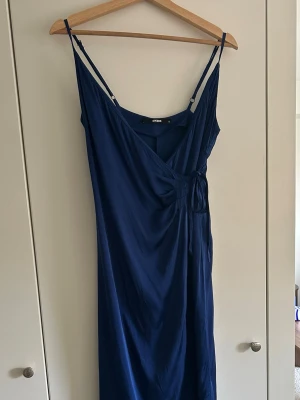 Marinblå omlottklänning från Bik Bok - Säljer en marinblå ankellångklänning från Bik Bok med smala axelband och omlottdetalj framtill. Klänningen har en enkel, elegant siluett och justerbara axelband.💙 säljer då den är för liten, endast använd en gång!