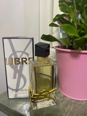 Yves Saint Laurent Libre Eau de Parfum - Yves Saint Laurent Libre Eau de Parfum i en elegant, rektangulär glasflaska med guldiga detaljer och svart asymmetrisk kork. Doften kommer i originalförpackning med vit och guld design. Perfekt för dig som gillar lyxiga och moderna parfymer.